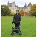 A person stands on a green lawn with the 15-lb Triumph Mobility Escape Rollator, featuring a one-handle fold and advanced brakes, in front of a historic building with towers amidst autumn-colored trees and cloudy skies.