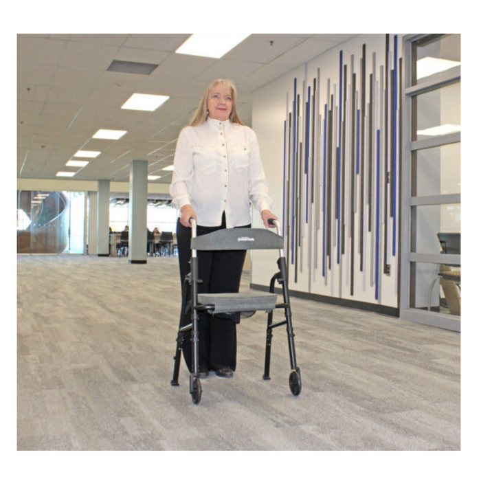 A woman with long gray hair and a white blouse uses the Triumph Mobility Essentials Walker with Seat in a spacious, well-lit office hallway. The walls have vertical stripes, and large windows are in the background.