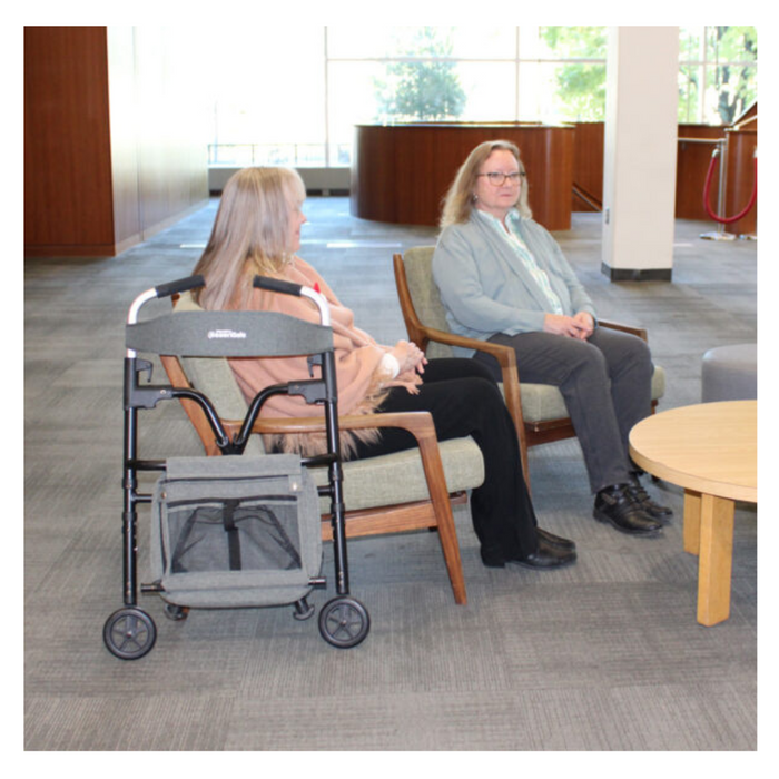 Two people are seated in a lounge area with large windows and wooden furniture, engaged in conversation. Nearby is a Triumph Mobility Essentials Walker with Seat, contributing to the relaxed and accessible environment.