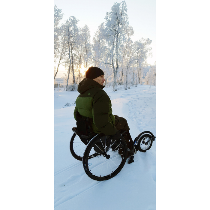 A person wearing a green jacket and black hat enjoys the winter landscape in an off-road wheelchair equipped with Triumph Mobility reTyre Traction Skins, ensuring excellent traction and puncture protection on a snowy path surrounded by snow-covered trees as the sun sets warmly.