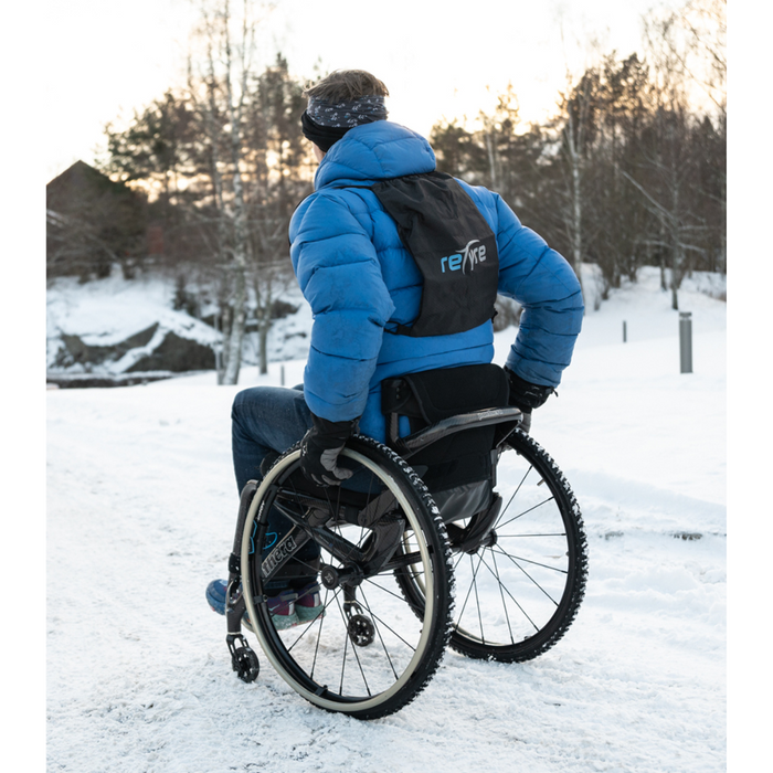 Wearing a blue jacket, gloves, and a black backpack, a person uses their wheelchair equipped with Triumph Mobility reTyre Traction Skins for exceptional traction on a snowy path surrounded by snow-covered trees and the setting sun.