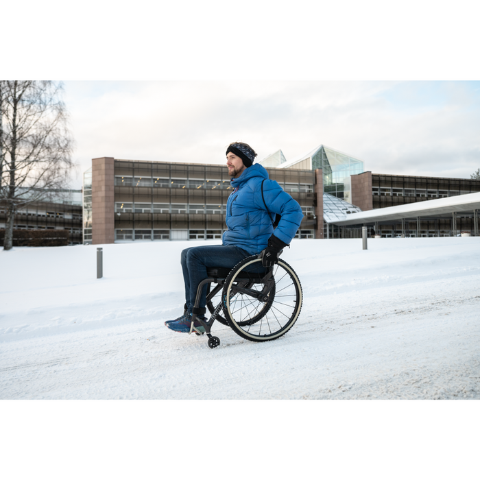 In front of a modern building, a person in a wheelchair expertly navigates a snowy path using Triumph Mobility reTyre Traction Skins. Theyre dressed in a blue jacket, gloves, and black beanie against a backdrop of cloudy skies and bare trees.