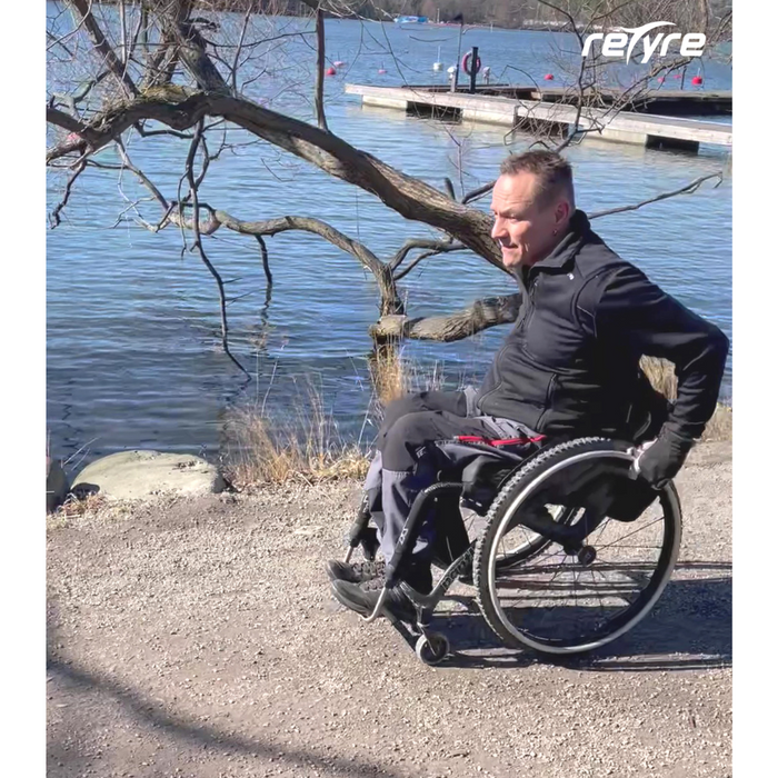 A person in a wheelchair with Triumph Mobility reTyre Traction Skins, featuring durable puncture protection, enjoys a sunny day by the lakeside path, with a leafless tree and dock in the background.