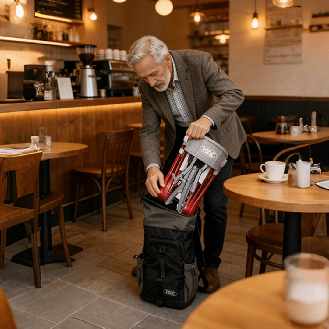 In a warmly lit, empty café surrounded by tables with cups and plates, an older man packs a folded Ultra-Compact PackGo Rollator Walker with Backpack (VOCIC Z35)—its red and silver frame fitting neatly—into a black bag.
