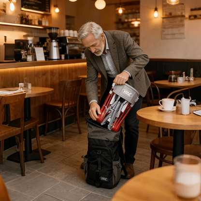In a warmly lit, empty café surrounded by tables with cups and plates, an older man packs a folded Ultra-Compact PackGo Rollator Walker with Backpack (VOCIC Z35)—its red and silver frame fitting neatly—into a black bag.