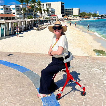 A woman in a sun hat sits on the Ultra-Compact PackGo Rollator Walker with Backpack | VOCIC Z35 near a beach, smiling. The background shows sand, people, palm trees, and resort buildings under a clear blue sky.