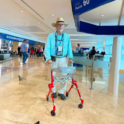An older man in a hat and blue shirt smiles while using a red Ultra-Compact PackGo Rollator Walker with Backpack | VOCIC Z35 at an airport near gate 60, with travelers and signs visible in the background.