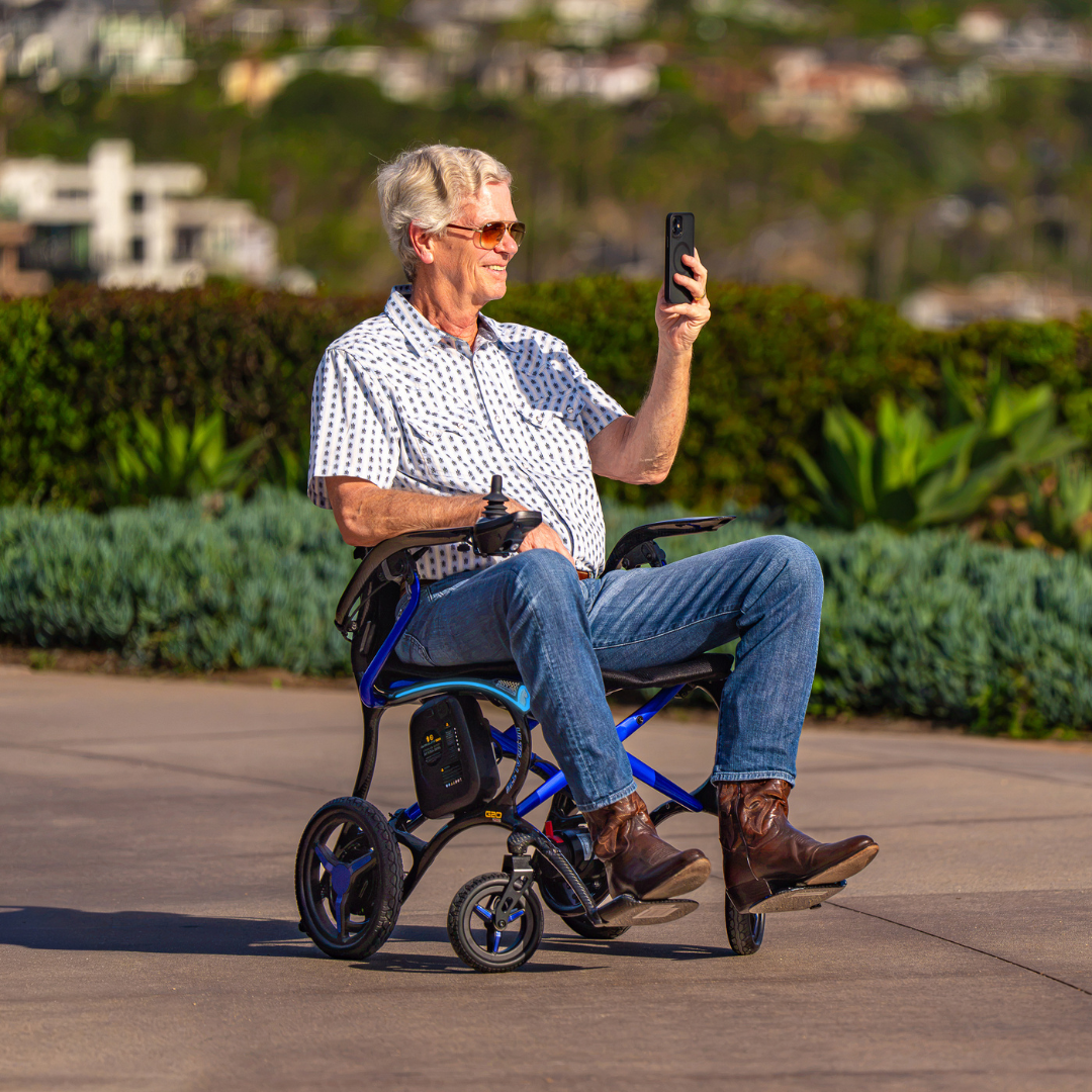 An older man in sunglasses sits outdoors in a Forcemech Ultralite G20 PRO Carbon-Fiber Power Wheelchair, smiling as he holds up a smartphone. He is dressed in a patterned shirt, jeans, and brown boots, with green shrubs and buildings visible in the background.
