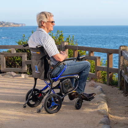 An older man in sunglasses sits in a Forcemech Ultralite G20 PRO Carbon-Fiber Power Wheelchair, enjoying an ocean view from a dirt path lined with a wooden fence and greenery under a clear blue sky.