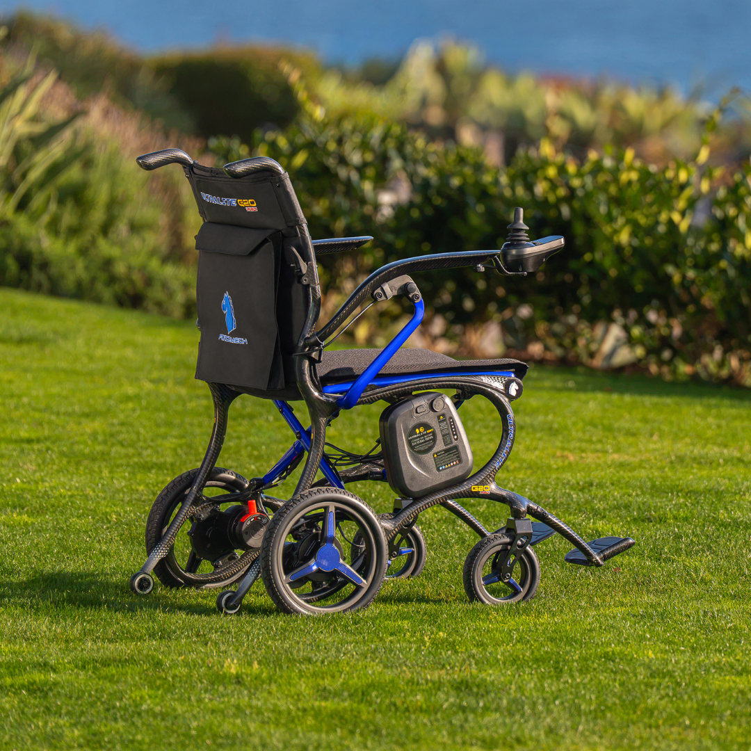 The Forcemech Ultralite G20 PRO Carbon-Fiber Power Wheelchair, featuring a black seat and an eye-catching blue frame, rests on green grass with trees and water softly blurred in the background. Its carbon-fiber construction is complemented by a visible control panel and joystick located on the armrest.