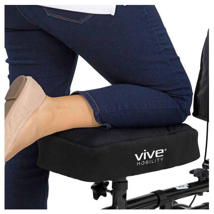 Wearing blue jeans and beige slip-on shoes, a person uses a knee scooter, their right knee resting on the Vive Mobility seat. Enhanced by the Vive Health Memory Foam Knee Walker Pad, it provides durable comfort against the plain white background.