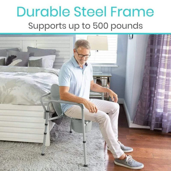 A man sits on a gray portable chair with armrests in a bright bedroom, its durable steel frame similar to the Vive Health Freestanding Bariatric Steel Bedside Commode supporting up to 500 pounds. Nearby, the room features a bed with gray and white bedding and a window with purple curtains.