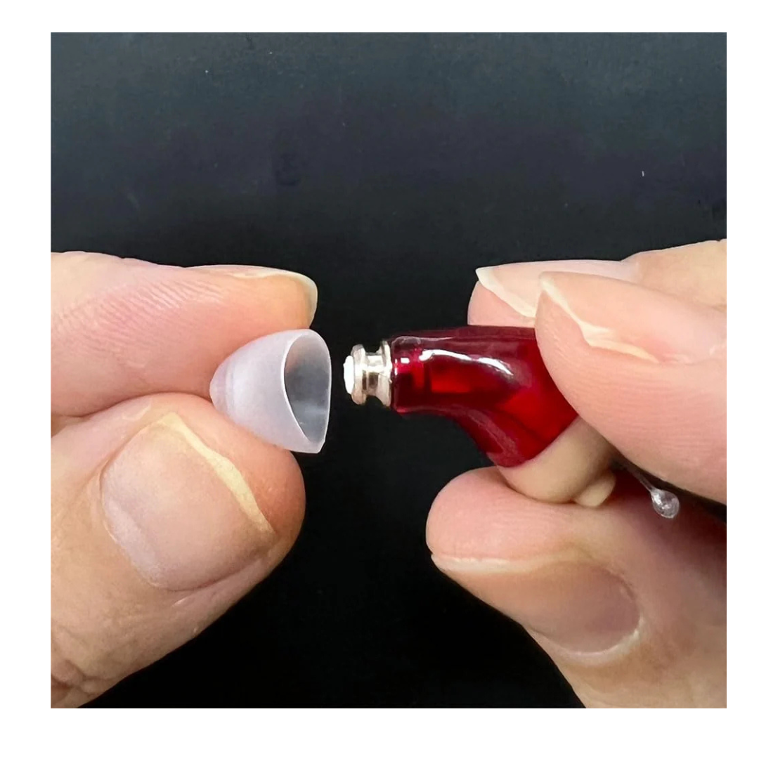 A close-up of two hands attaching a Vivtone Ear Tip Replacement for Super-Mini Hearing Aids to a small red in-ear device against a black background, highlighting essential accessory options.