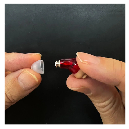 A close-up of two hands holding Vivtone Ear Wax Guards For Hearing Aids accessories—a small red device and a translucent silicone ear tip—against a black background.
