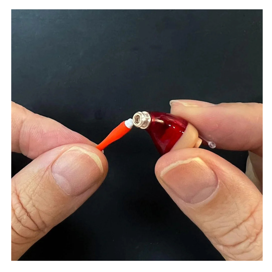 Two hands hold a Vivtone Ear Wax Guard for Hearing Aids, attaching or removing an orange rod-like piece on a small red and beige figure with a silver tip against a black background.