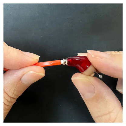 Close-up of hands using an orange tool to attach a red Vivtone Ear Wax Guard for Hearing Aids with a silver screw against a black background.