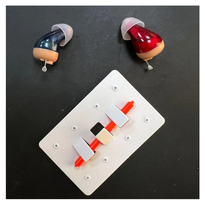 Two custom in-ear hearing aids, one blue and one red, are placed above a white rectangular electronic component—alongside Vivtone Ear Wax Guards For Hearing Aids and other accessories—on a dark surface.