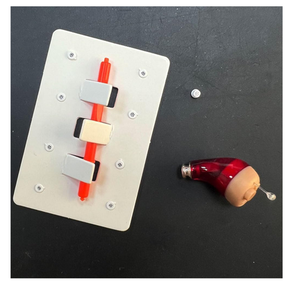 A white rectangular panel with three switches and a red slider sits on a dark surface next to a red and beige hearing aid, a small round battery, and Vivtone Ear Wax Guards For Hearing Aids.