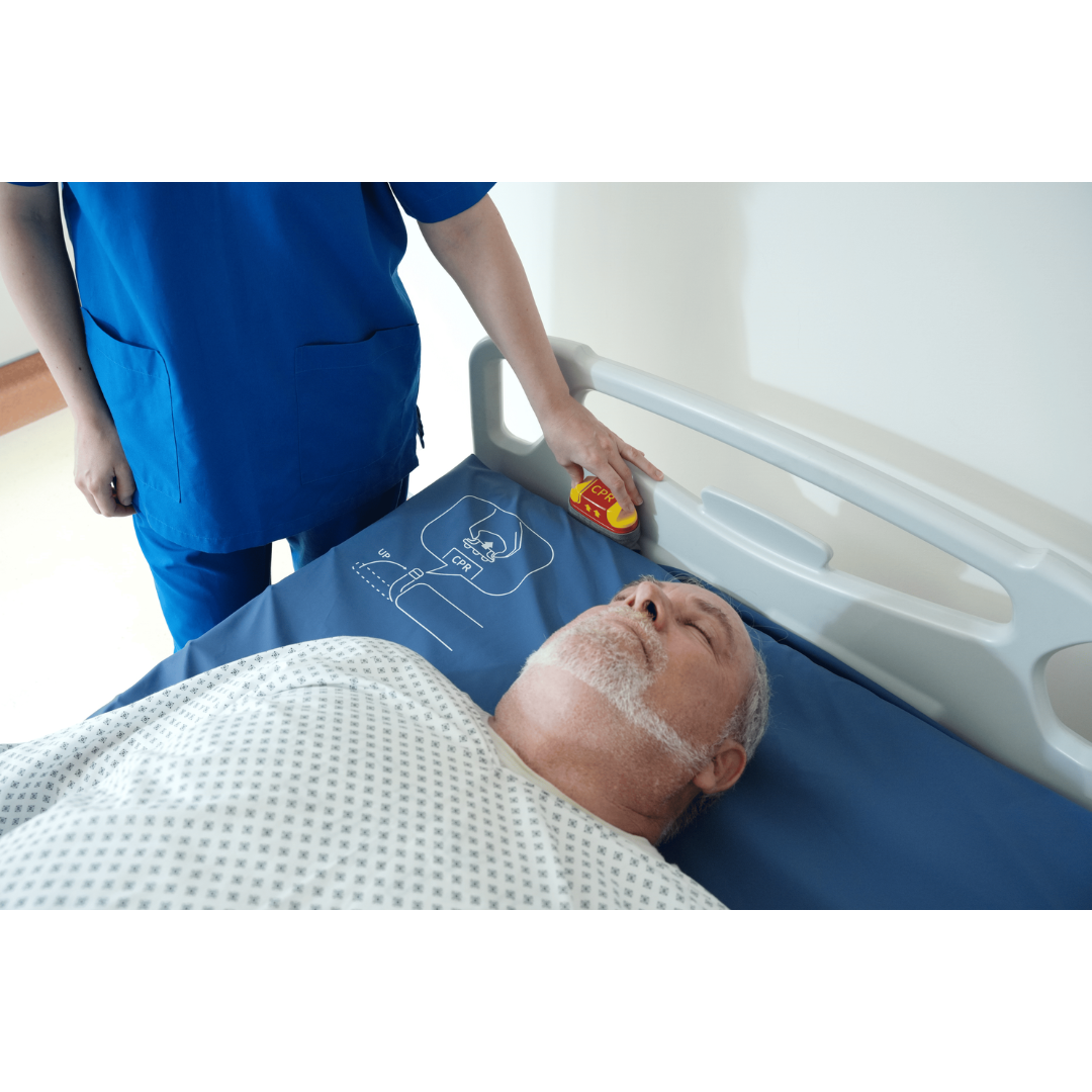 A nurse in blue scrubs stands by a hospital bed, holding a device near the head of an older male patient lying down with his eyes closed, covered by a patterned hospital blanket and resting comfortably on a Wellell Procare Auto G3 Hospital Air Mattress System.