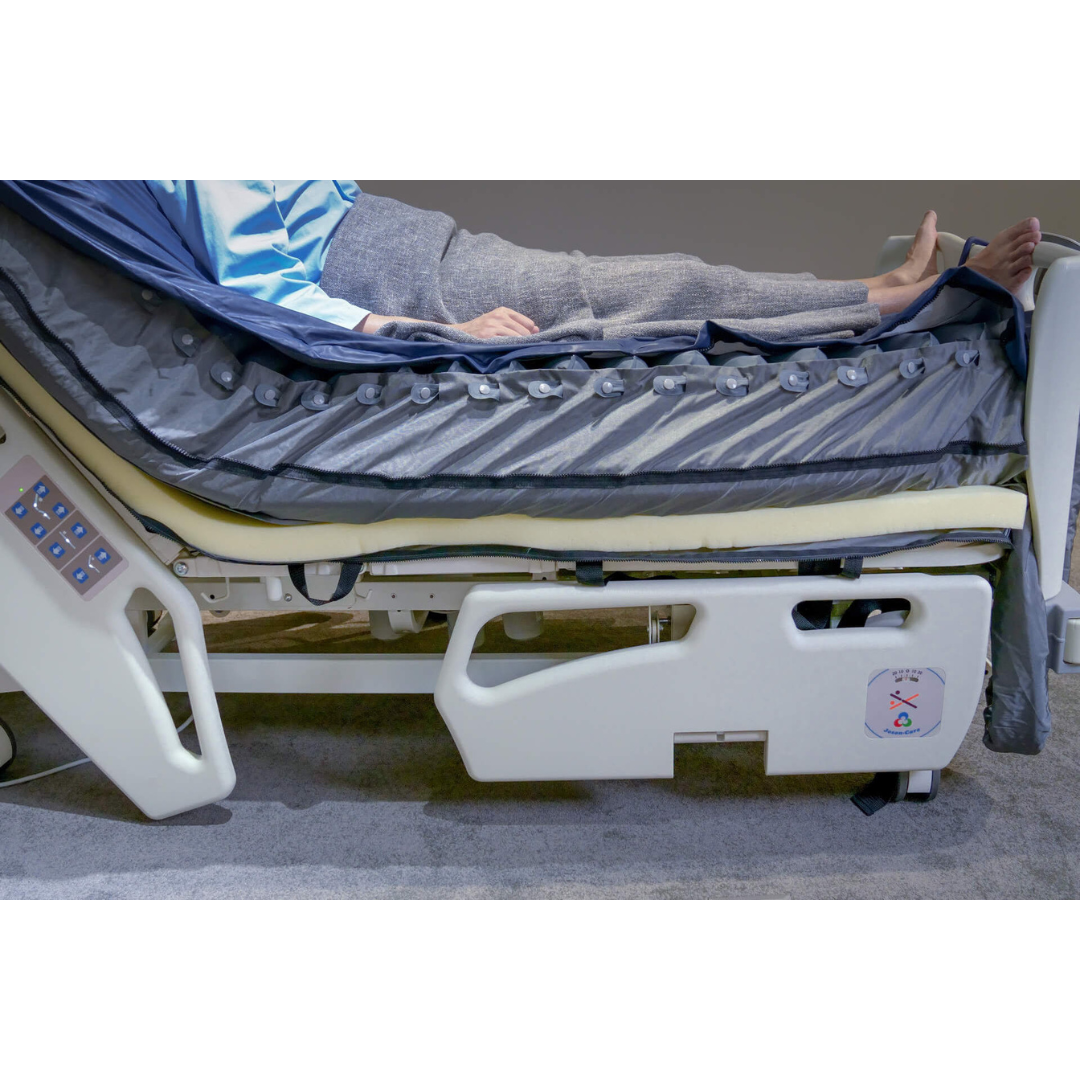 A person lies in a hospital bed, partially covered by a gray blanket. The Wellell Serene Air Alternating Pressure Mattress with True Low Air Loss features control panels and safety labels to help prevent pressure injuries.
