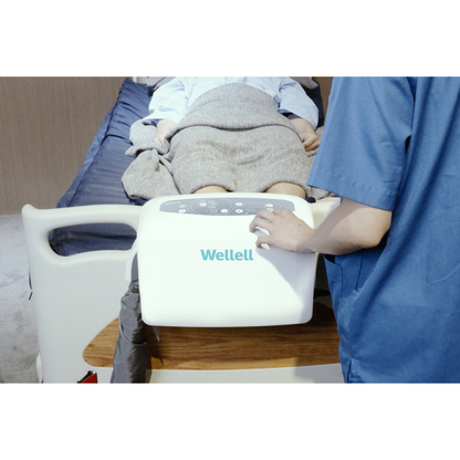 A healthcare worker in blue scrubs uses the Wellell Serene Air Alternating Pressure Mattress with True Low Air Loss beside a patient in bed, helping prevent pressure injuries and enhance comfort during post-acute care.