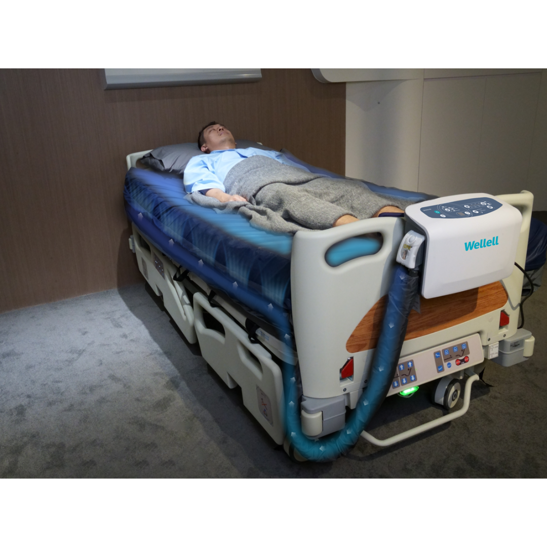 A person lies in a hospital bed, covered with a gray blanket. The bed features the Wellell Serene Air Alternating Pressure Mattress with True Low Air Loss, equipped with controls, medical devices, and a blue air hose labeled Wellell.