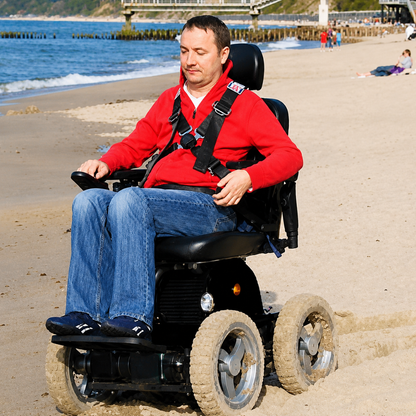 A man in a red jacket and blue jeans rides a Wheelchair88 All-Terrain Stair Climbing 4x4 Recreational Electric Vehicle on a sandy beach, with the sea and a pier visible in the background.