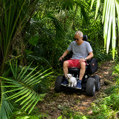 A man rides the Wheelchair88 All-Terrain Stair Climbing 4x4 Recreational Electric Vehicle along a forest trail, surrounded by lush greenery, with a small white dog on his lap.