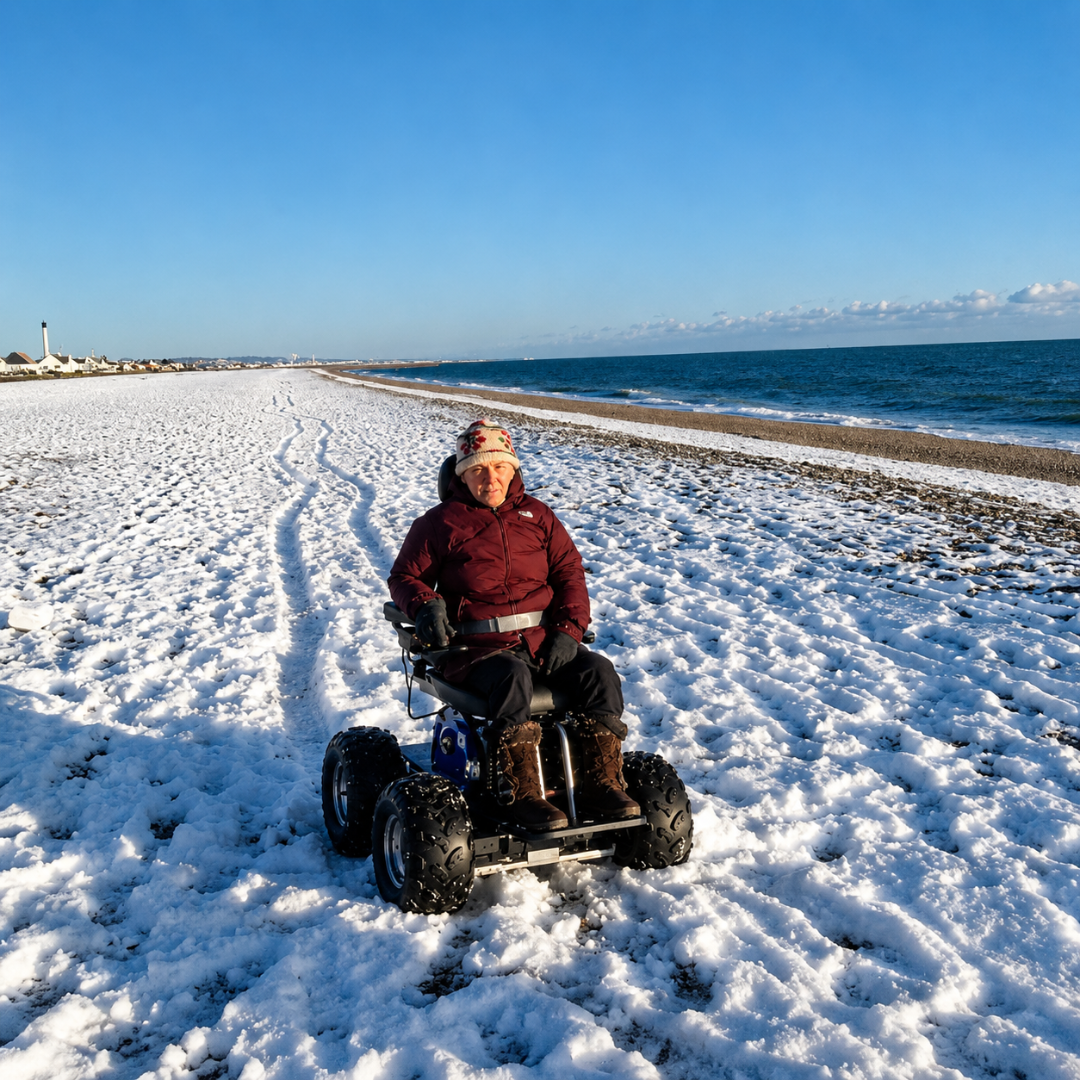 A person in a red jacket and winter hat uses the Wheelchair88 All-Terrain Stair Climbing 4x4 Recreational Electric Vehicle on a snow-covered beach by the ocean under a clear blue sky.