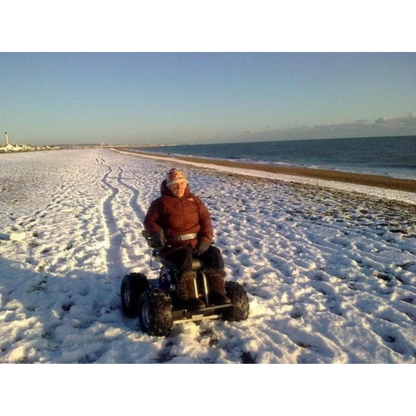 A person in a wheelchair with large off-road wheels travels along a snowy beach near the shore in a Wheelchair88 All-Terrain Stair Climbing 4x4 Recreational Electric Vehicle, leaving tracks in the snow with the sea and blue sky behind them.