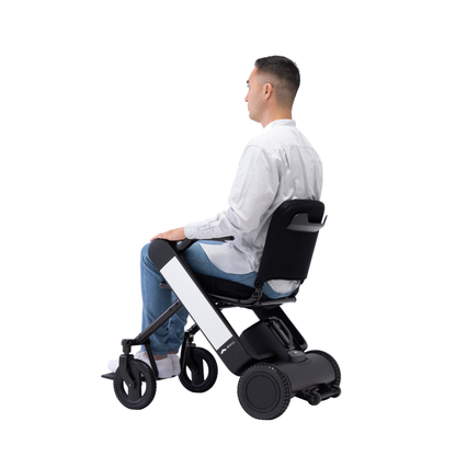 A man in a white shirt and light blue jeans sits in a Whill Model F Lightweight Portable Electric Wheelchair, facing right against a plain white background. Warranty details and specifications available upon request.