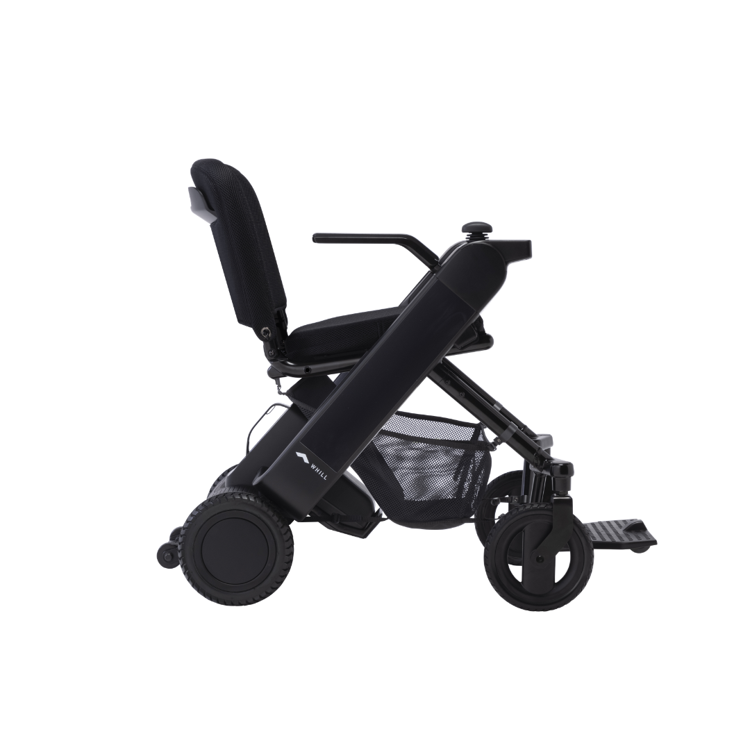 The Whill Model F Lightweight Portable Electric Wheelchair is shown from the side on a white background, featuring a cushioned seat, backrest, armrests, mesh storage basket, and large rear wheels. View specifications and warranty details for more info.