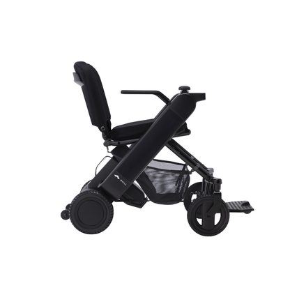 The Whill Model F Lightweight Portable Electric Wheelchair is shown from the side on a white background, featuring a cushioned seat, backrest, armrests, mesh storage basket, and large rear wheels. View specifications and warranty details for more info.