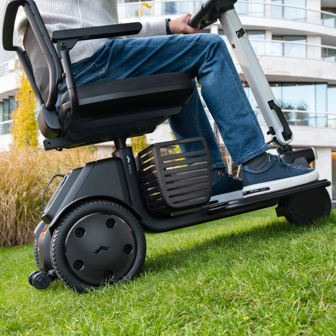 A person in jeans and sneakers rides the Whill Model R 3-Wheel Mobility Scooter across grass in an outdoor urban area with apartment buildings visible in the background.