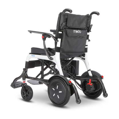 The Whill MODO Breeze Ultra-Lightweight CF Folding Electric Wheelchair is a modern powerchair featuring a black seat and backrest, a meddo-labeled storage pouch, and a white frame with black accents and large rear wheels. Shown from the rear, this ultra-lightweight electric wheelchair combines style with convenient portability.