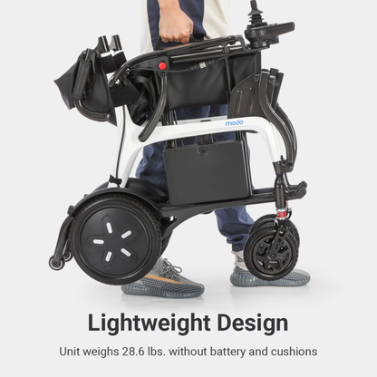 A person stands holding a compact, folded Whill MODO Breeze Ultra-Lightweight CF Folding Electric Wheelchair by the frame. This foldable powerchair features large rear wheels, small front wheels, and a sleek black-and-white design. Text highlights its ultra-light weight and convenient folding design, making it easy to transport and store.
