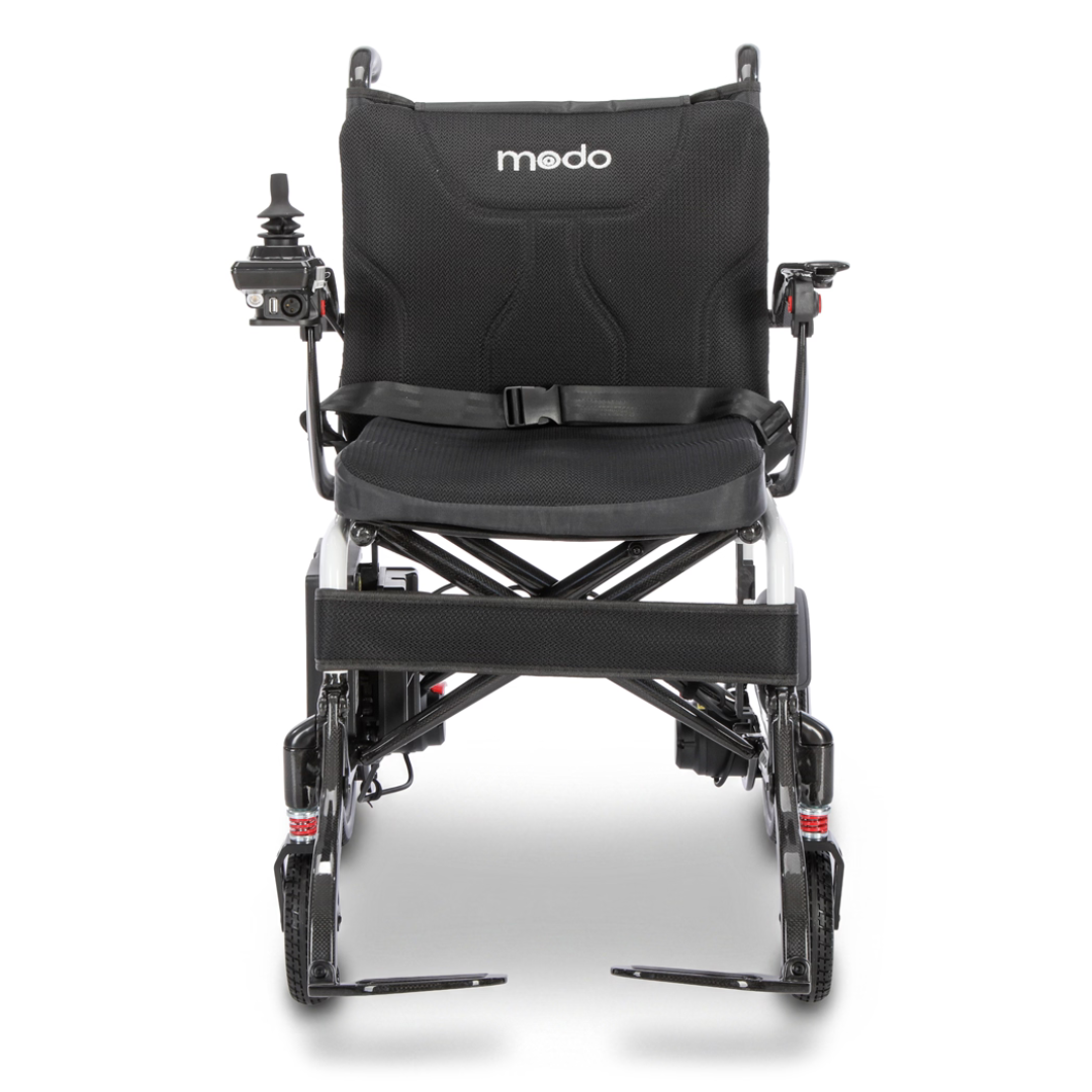 Front view of a black Whill MODO Breeze Ultra-Lightweight CF Folding Electric Wheelchair featuring modo branding on the backrest, joystick control on the left armrest, seatbelt, and footrests.