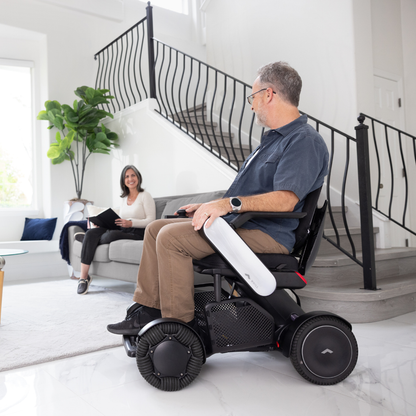 A man in a Whill Model C2 Smart Power Chair faces a smiling woman on a couch in a bright, modern living room with a large staircase and potted plant.
