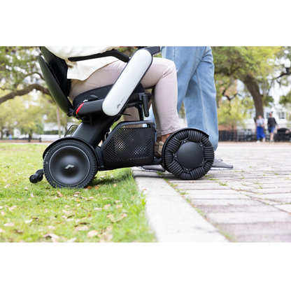 A person in a Whill Model C2 Smart Power Chair - Compact Portable Mobility approaches a curb, while another stands nearby on a paved path in a park with green trees in the background.