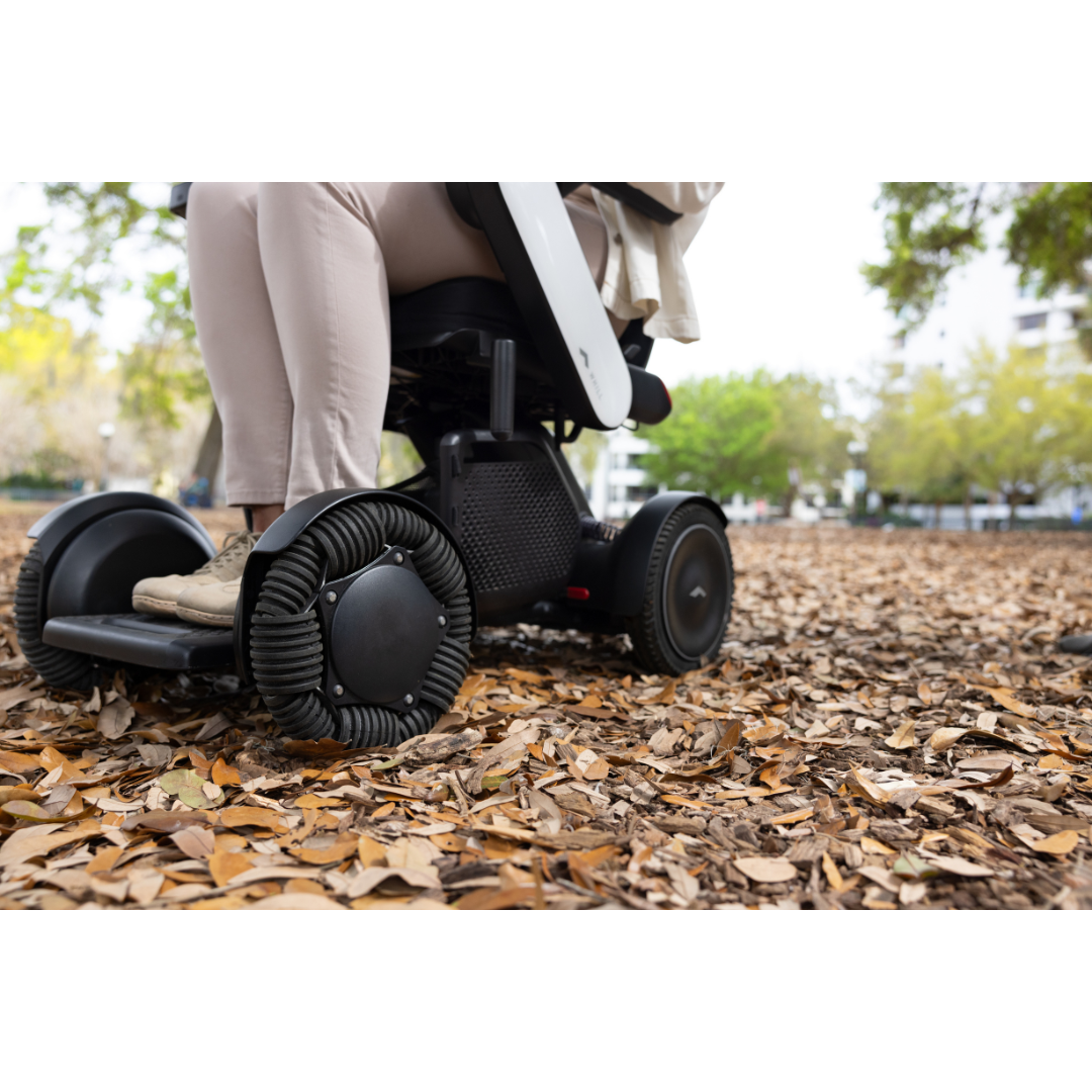 A person uses the Whill Model C2 Smart Power Chair - Compact Portable Mobility, which features unique treaded wheels, to navigate over dry leaves in an outdoor park.