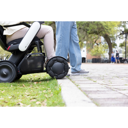 A person in a Whill Model C2 Smart Power Chair - Compact Portable Mobility with advanced wheels is next to another person standing on a park sidewalk, with trees and people visible in the background.
