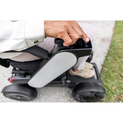 A person’s hand operates the control panel of a Whill Model C2 Smart Power Chair on a paved path next to grass, with their legs and part of this compact, portable mobility device visible.
