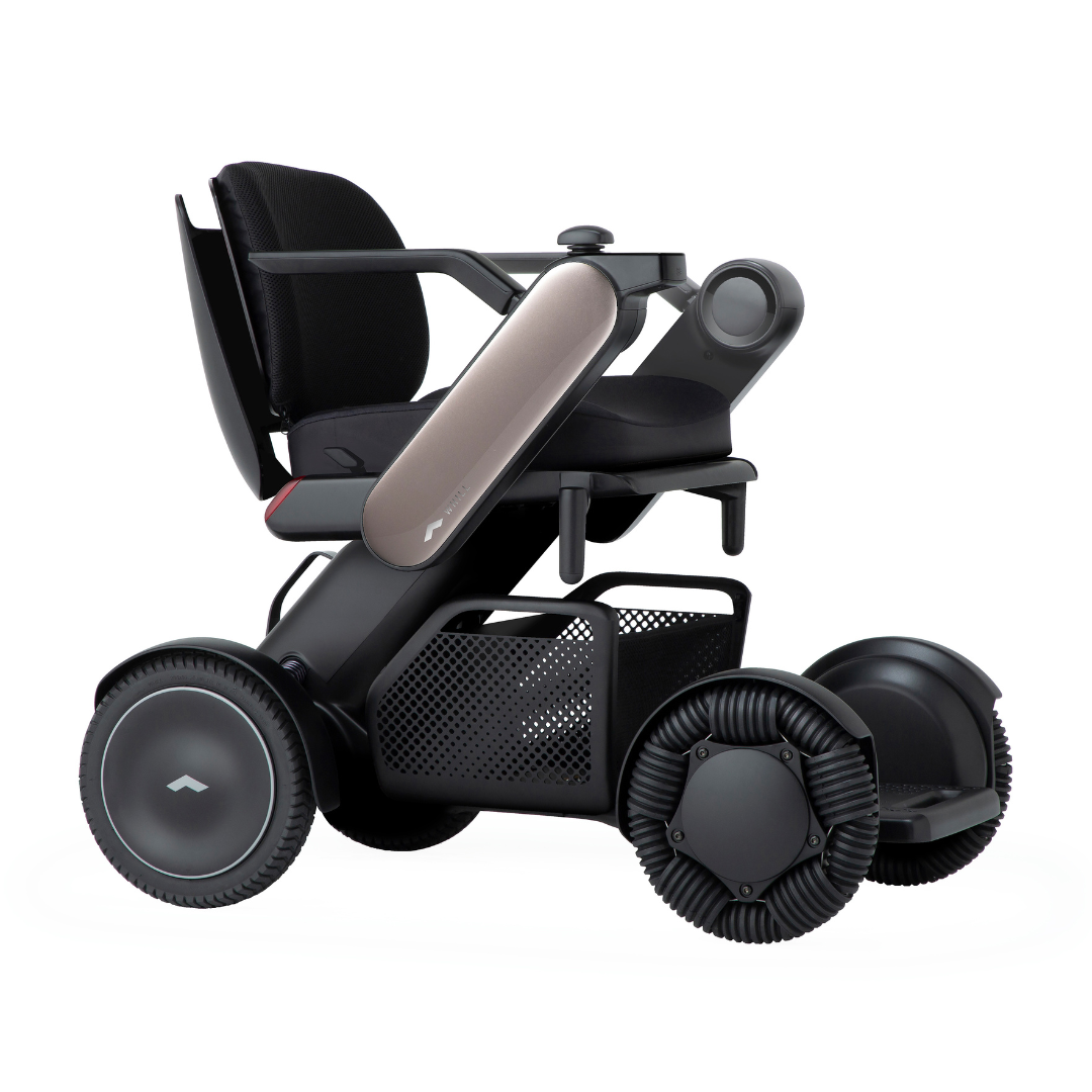The Whill Model C2 Smart Power Chair - Compact Portable Mobility features a sleek black and metallic design, large wheels, padded seat, mesh storage basket, and futuristic armrests. It’s a Class II medical device with modern, minimalist style.