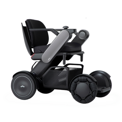 The Whill Model C2 Smart Power Chair is a compact, portable mobility device in black with large wheels, a mesh basket under the seat, contoured armrests, and minimalistic design—engineered as an FDA-compliant Class II medical device.