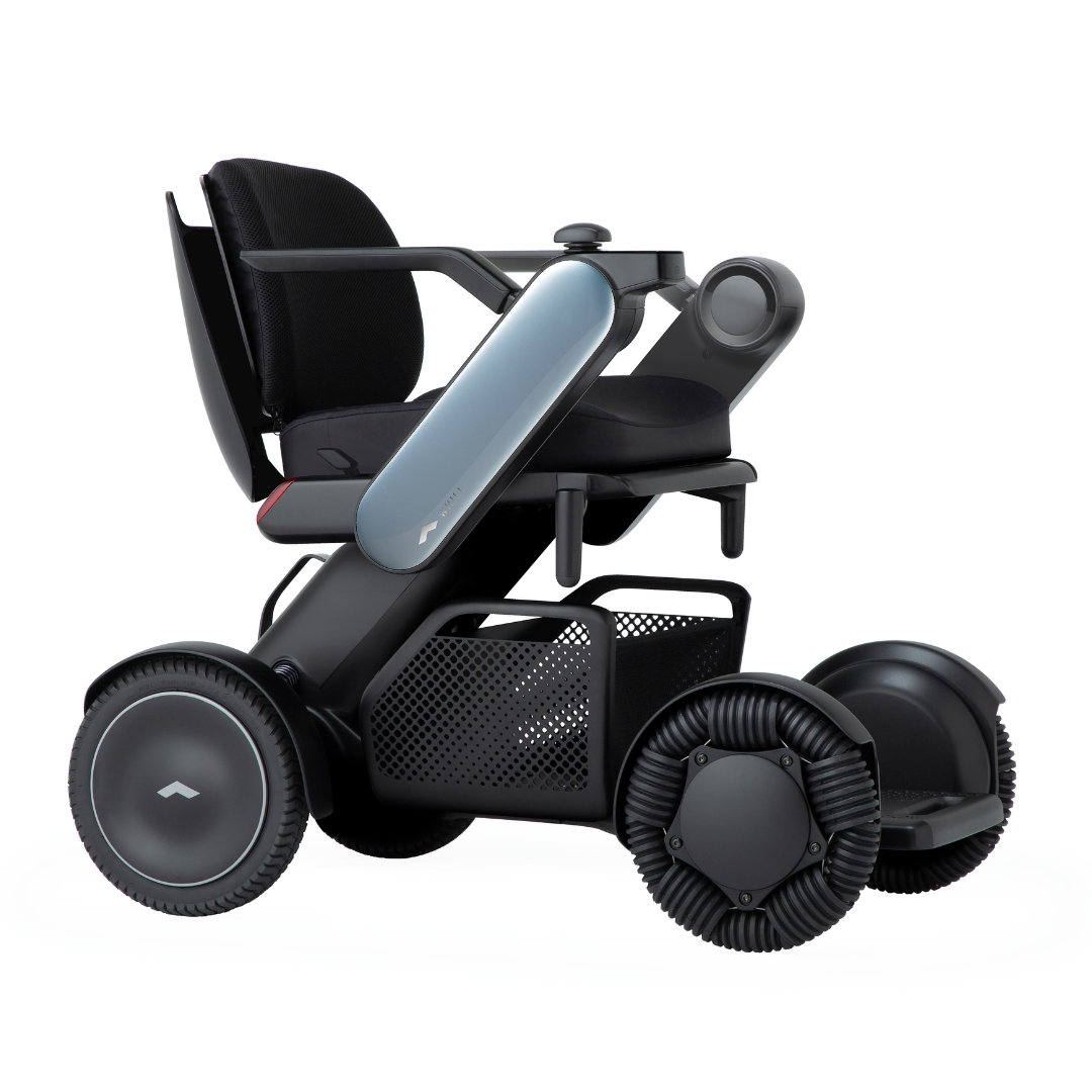 The Whill Model C2 Smart Power Chair is a compact, portable electric wheelchair with large all-terrain wheels, a black cushioned seat, armrests, storage basket, and a sleek minimalist design. It’s a Class II medical device with smooth, futuristic styling.