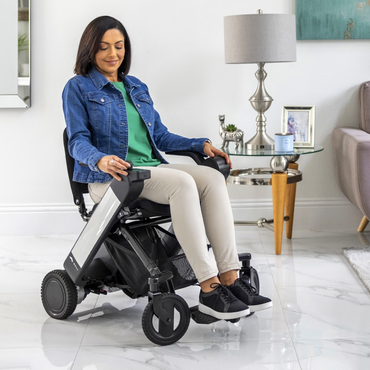 A woman relaxes in a Whill Model F Lightweight Portable Electric Wheelchair in a bright, stylish living room, reviewing its specifications and warranty details on her tablet near a side table with framed photos.