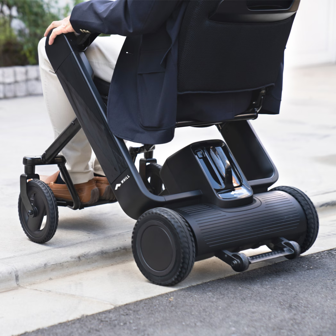 A person in business attire uses the Whill Model F Lightweight Portable Electric Wheelchair with large wheels, designed for easy transitions from sidewalk to street while meeting the latest specifications.