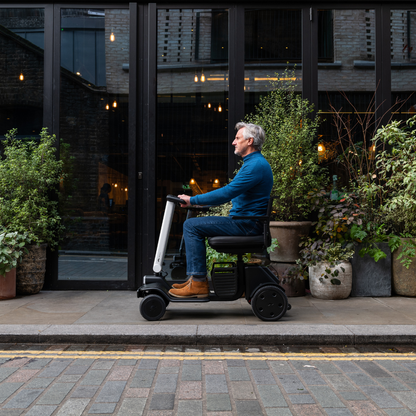 A man in a blue sweater and brown boots rides the Whill Model R 4 Wheel Portable Scooter along a city sidewalk past potted plants by a glass building, showcasing its convenience and accessibility for daily urban mobility.