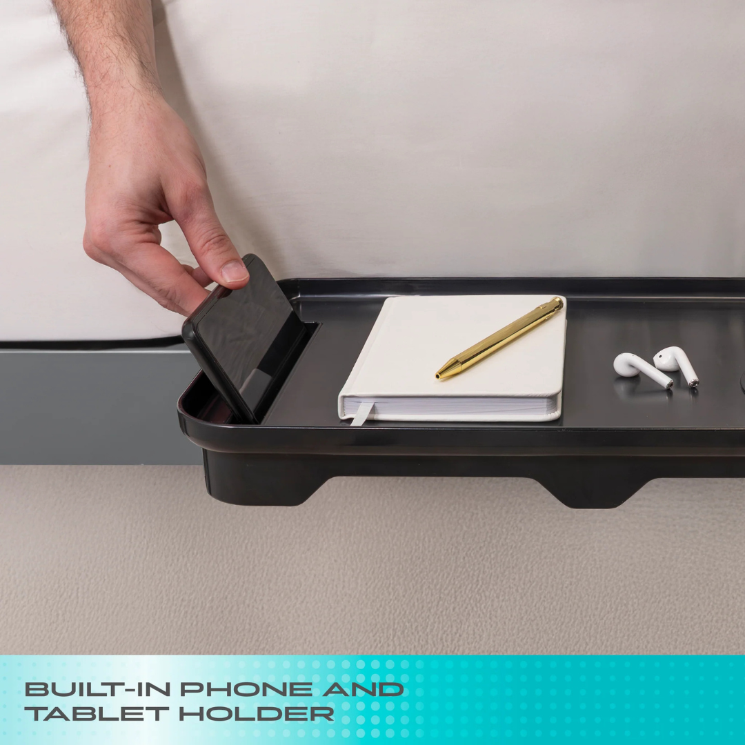 A hand places a smartphone into the built-in holder of the black Stander Wonder Bedside Shelf Tray, alongside a notebook, pen, and wireless earbuds—offering a smart space-saving solution. Text reads: BUILT-IN PHONE AND TABLET HOLDER.