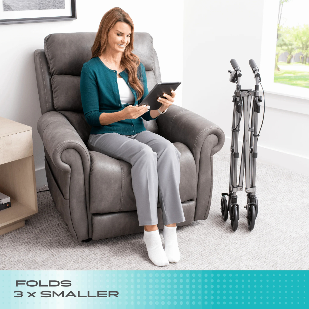 A woman reads a tablet in a gray recliner, with the Stander Wonder Indoor Rollator with Tray Table folded nearby. Text at the bottom says, FOLDS 3x SMALLER. Bright room and large window in the background.